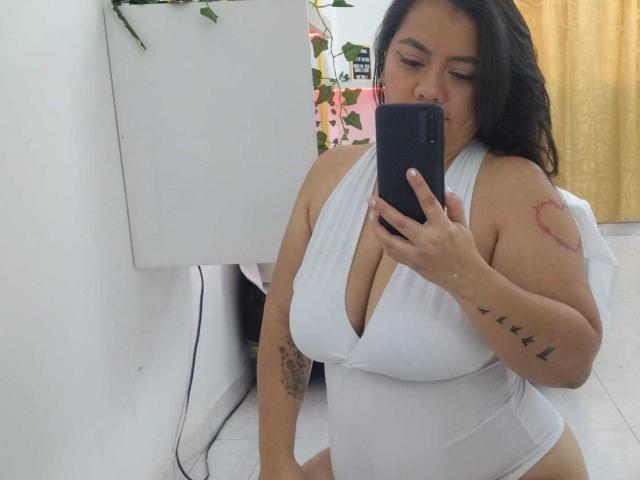 Femme pulpeuse et voluptueuse, avec de grandes seins, portant une combinaison blanche à encolure dégagée, se prenant en photo dans un miroir, tenant un téléphone noir, montrant des tatouages sur le bras et le côté, cheveux bruns..