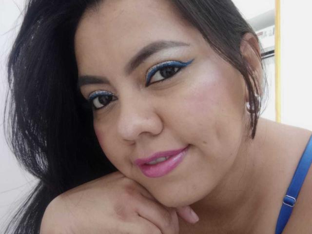 Femme corpulente et pulpeuse, avec des cheveux foncés, un eye-liner bleu, du rouge à lèvres rose, souriante, le menton appuyé sur la main, un bandeau bleu visible, des boucles d'oreilles de perles, une peau claire, des joues légèrement rosées, regardant la caméra..
