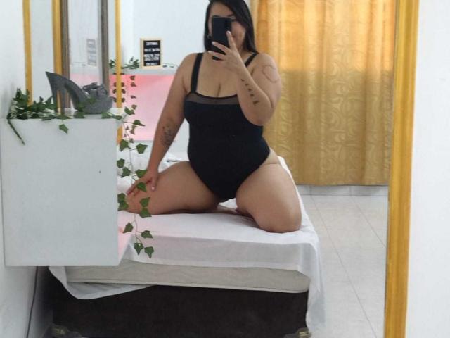 Femme pulpeuse et voluptueuse en maillot de bain noir une pièce, à genoux sur le lit, montrant ses seins et ses fesses, tenant un téléphone, selfie devant un miroir, tatouages sur le bras, cheveux bruns, rideau jaune en arrière-plan..