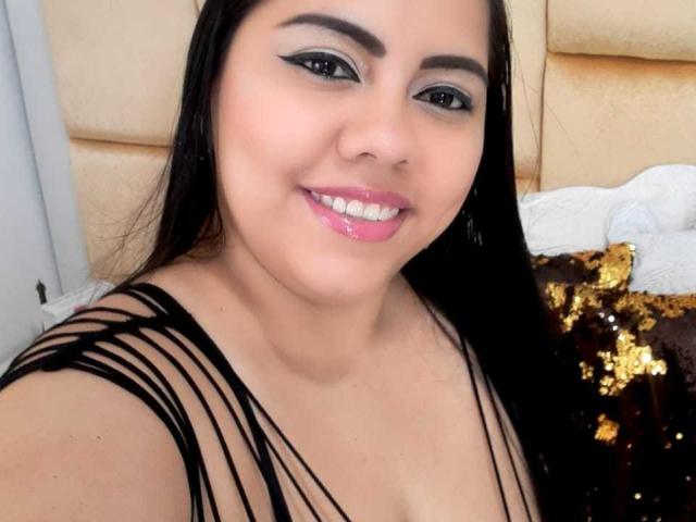Une femme corpulente et voluptueuse, avec des cheveux noirs, des lèvres roses et brillantes, une robe noire à bretelles, et un eye-liner bleu, souriant à la caméra, montrant légèrement ses seins. Elle a les yeux marron, une peau claire et est mignonne..
