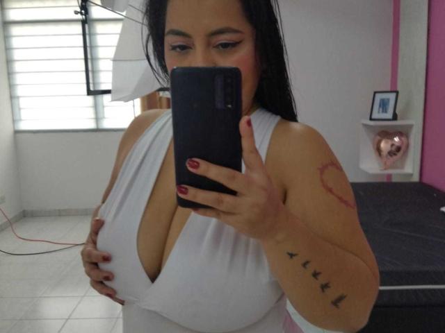 ChubbyThickMilf with black hair, white deep cleavage dress, red nail polish, holding phone, tattoo on arm, looking down, in a room with pink wall and white window.
