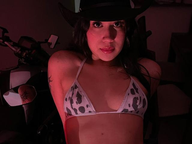 HannaThorne portant un haut de bikini à motifs de vache, un chapeau noir, avec ses seins visibles, regardant vers le haut avec une peau rougeoyante, et un tatouage sur son bras droit..