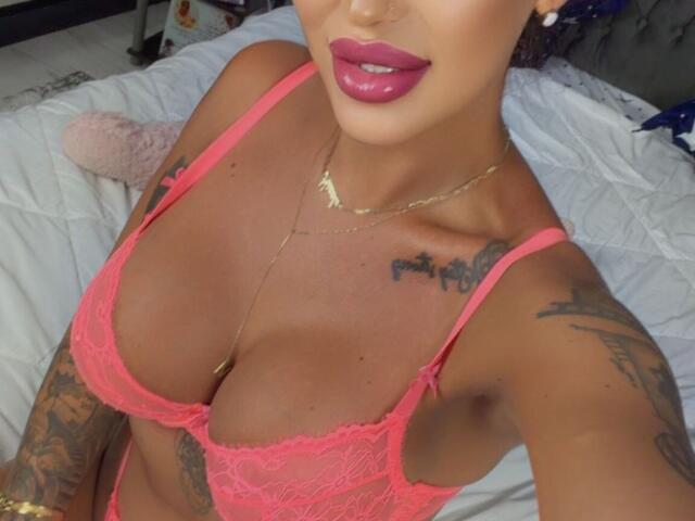 NicolSNaya, avec une peau bronzée, un soutien-gorge en dentelle rose, de grandes seins, des tatouages sur les bras, un rouge à lèvres rose foncé, un collier en or, allongée sur un lit, prenant un selfie, montrant sa poitrine, souriante, regardant la caméra..