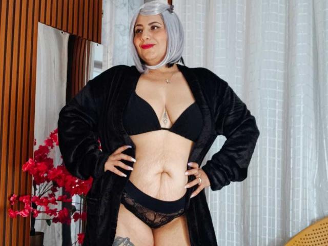 AngeleRouxo stands with hands on hips, wearing a black bra and lace panties, black robe open, grey bob, red lipstick, choker, tattoo below boobs, gray hair, confident smile.