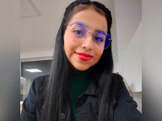 EmmaSollins has long black hair, blue and purple transparent glasses, red lipstick, and a black jacket over a green turtleneck. She's smiling with a cute nose ring.