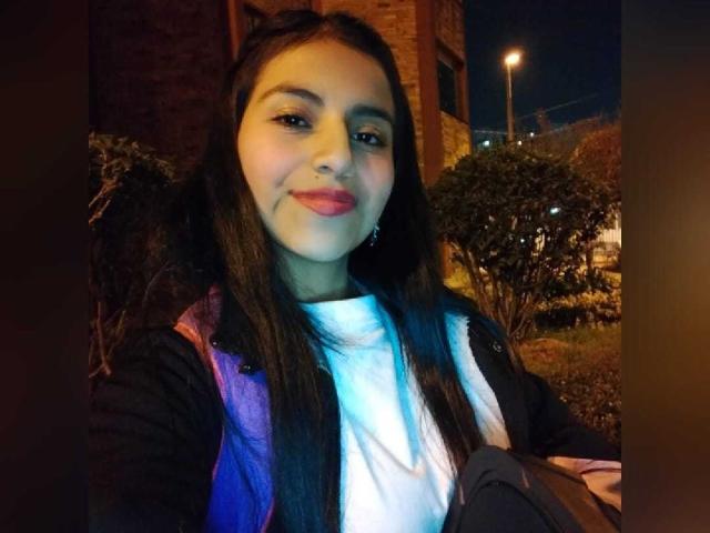 EmmaSollins with long black hair, pink lipstick, white shirt, black jacket, purple backpack, and earrings, smiling at night, streetlights in background, holding gray bag.