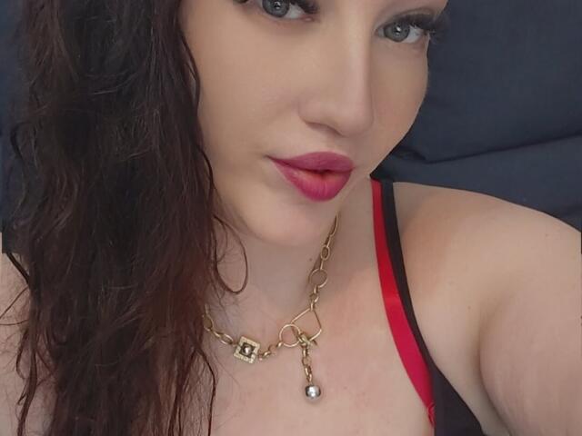 VanessaSquirtt has light skin, wet brown hair, and red lipstick. She's wearing a gold chain necklace with a pearl and a red-black top. Her tits are slightly visible.