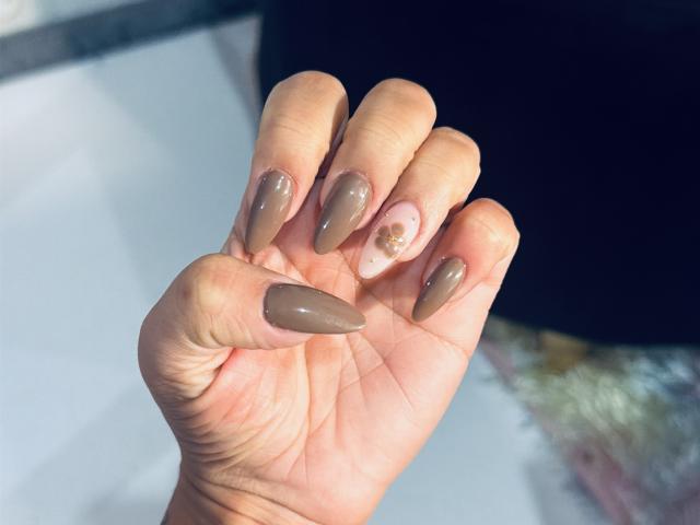 A mão de ThaliaAres, com unhas acrílicas longas, brilhantes e marrons, e uma unha rosa com um design de glitter dourado, exibindo suas pontas finas e brilhantes..