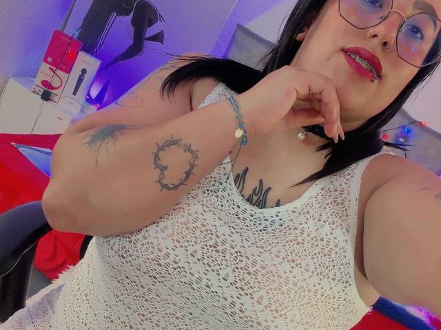 NatalieHarris with black hair, glasses, red lipstick, white lace sleeveless top, showing tattoos on her arm and chest, finger near mouth, blue bracelet, and black choker.