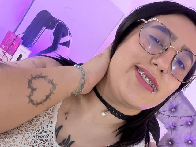 NatalieHarris with glasses, black hair, nose ring, and tooth jewelry, showing a crown tattoo on her hand, wearing a black choker and lace top, smiling. Purple room.