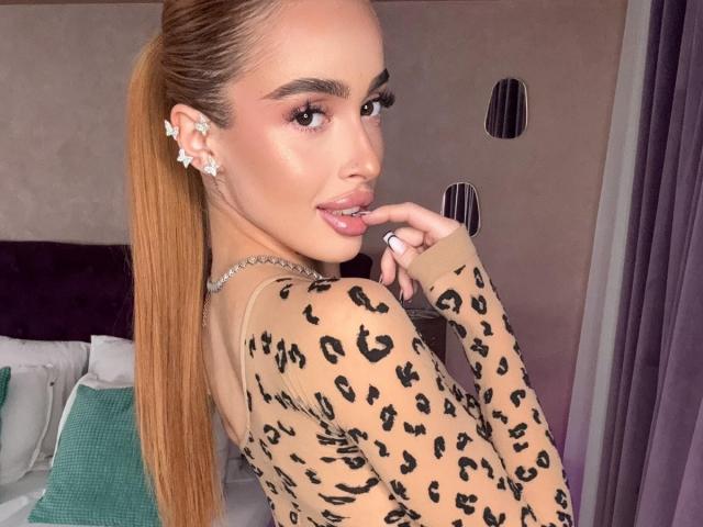 MarissaFex, con una larga coleta rubia, vistiendo una blusa de estampado de leopardo, mordiéndose el dedo, mostrando unos labios rosados brillantes y pendientes de diamantes, mirando hacia atrás con una sonrisa sensual..
