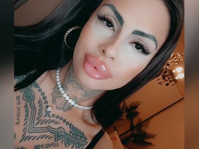 DenyChantal with black hair, full lips, and tattoos on her chest, wearing a diamond necklace and hoop earrings, sticking out her tongue in a selfie.