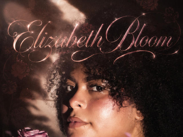 ElizabethBloom's face with curly black hair, shiny brown skin, and subtle makeup, looking seductively at the camera, with a pink floral accent in the lower left.