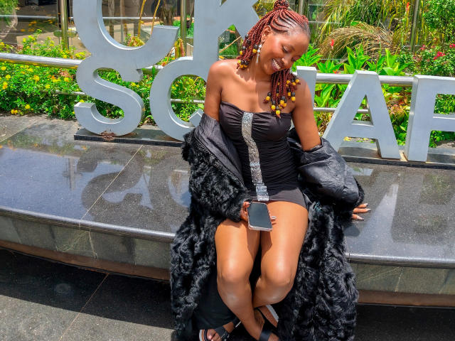 MonaX69 with brown skin, small tits, and braided red hair, sits on a stone bench, wearing a black strapless dress, furry coat, and holding a phone, smiling.