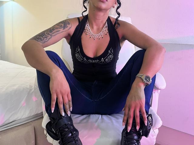 TracyLambo sits with legs spread, wearing a black halter top, blue jeans, and black boots. She has a tattooed arm, necklace, and watch, with visible boobs.