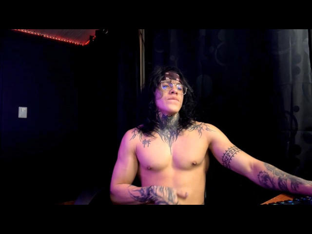 Shirtless KenshinOpt with tattoos, glasses, and long dark hair, sitting in a dimly lit room, arms resting on a surface, looking slightly upward with a calm expression.