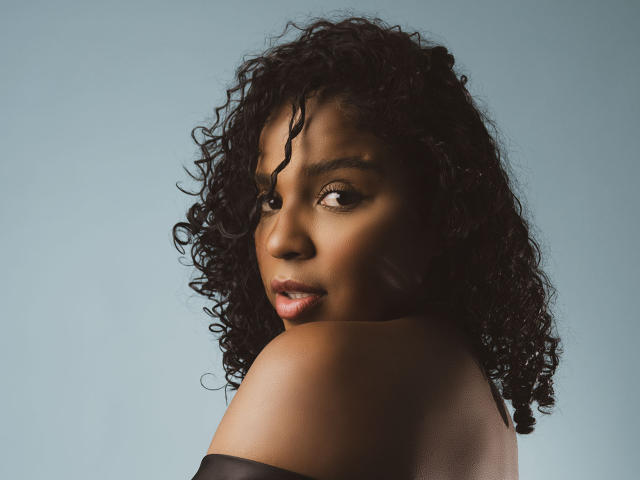 LisaMendez has dark curly hair, looking over her shoulder with a serious expression. Her bare shoulders and black off-shoulder top are visible. Her brown skin is smooth.