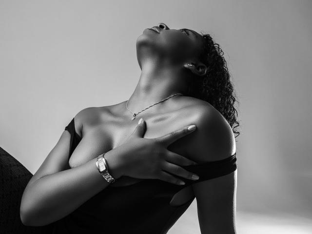 LisaMendez with dark skin, curly hair, and a necklace, head tilted back, covering her tits with one hand, wearing a black off-shoulder top, in a black-and-white photo.