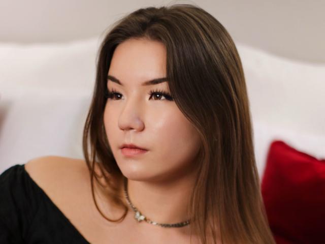 Iris Prescott tiene el cabello castaño lacio, piel clara y lleva una blusa negra con un hombro descubierto y un collar. Su expresión es neutra, con un maquillaje sutil y pestañas largas..