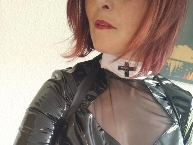 MissShine-hot in black shiny latex top with sheer mesh, white collar, and black cross, showing her upper chest and red lipstick. Her pinkish-red hair is shoulder-length.