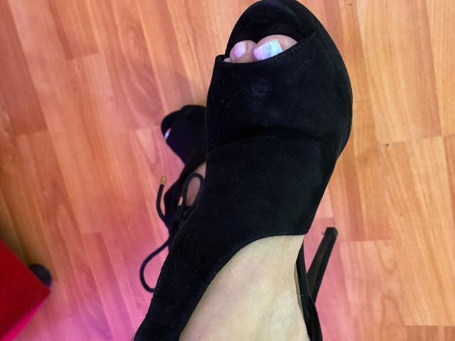 Karimelee's black high heel with an open toe, showing her white-painted toenails, is on a wooden floor, with the heel slightly tilted and the lace partially visible.