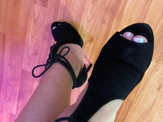 Karimelee's feet in black open-toe high heels with black straps, showing toes with pink nail polish, on a wooden floor.