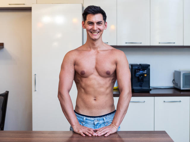 Clint Masters zonder shirt, met een gespierd en getraind lichaam, lachend, alleen gekleed in blauwe jeans met een zichtbare "Calvin Klein" tailleband, handen op een houten aanrecht..