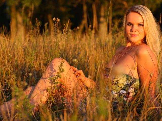 PenelopaMurMiay with blonde hair, sitting in tall grass, wearing a strapless dress, showing her tits, smiling, sunlight on her face, hand on knee, forest background.