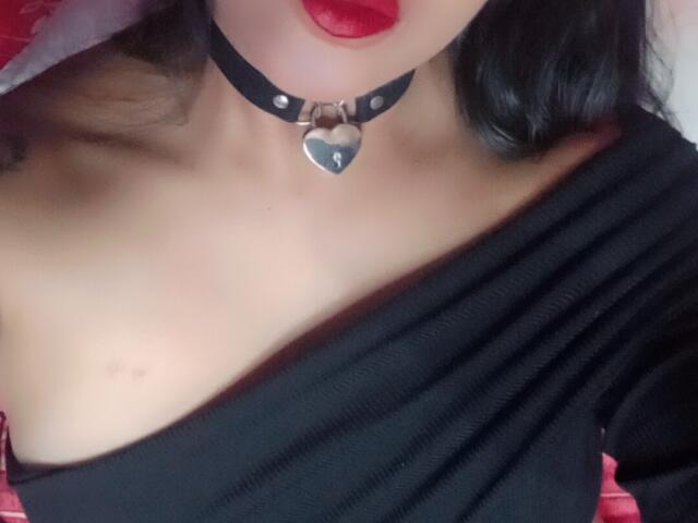 AmmyShtones with red lips, black choker, heart-shaped metal ring, and black off-shoulder top, showing a small, subtle tan line on her right boob.