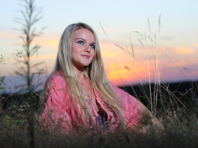 PenelopaMurMiay with long blonde hair, pink sheer top, and visible cleavage, sitting in tall grass at sunset, looking to the side with a slight smile.