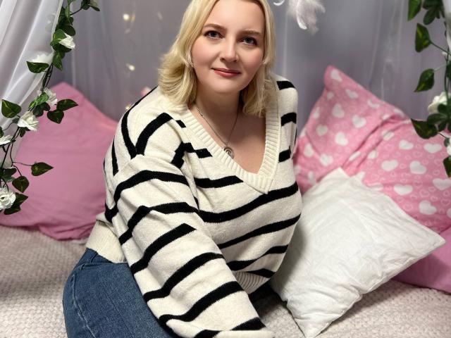 VictoriaNotti, blonde, smiling, wearing a black and white striped sweater, sitting on a bed with pink and white pillows, blue jeans, and white feather earrings.