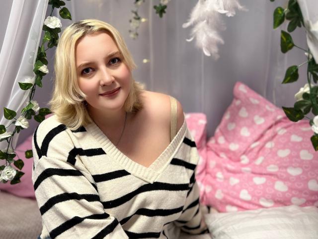 VictoriaNotti with blonde hair, wearing a black and white striped sweater, showing a small cleavage, smiling, sitting on a pink bed with heart-patterned pillows.