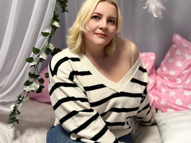 VictoriaNotti with blonde hair, wearing a black-and-white striped sweater, showing her tits through the V-neck, sitting on a bed with pink heart-patterned pillows.