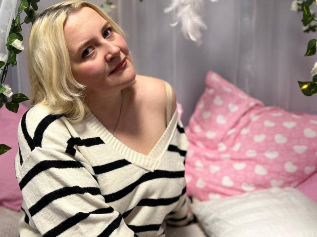 VictoriaNotti with blonde hair, off-shoulder striped sweater, and cute smile, showing her tits slightly, sitting on pink heart-patterned pillow, surrounded by green leaves.