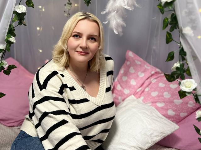 VictoriaNotti with blonde hair, wearing a black and white striped sweater, sitting on pink pillows with white heart patterns, smiling at the camera, her tits slightly visible.