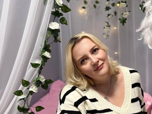 VictoriaNotti with blonde hair, smiling, wearing a black and white striped sweater, has a cute nose ring, and is looking slightly to the right, in a soft-lit setting.