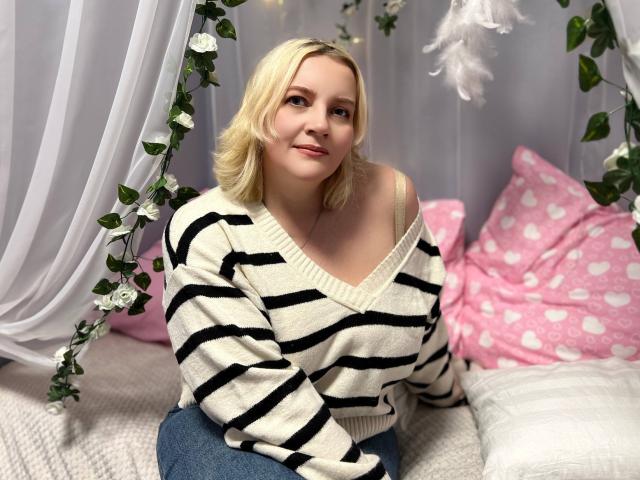 VictoriaNotti with blonde hair, off-shoulder black and white striped sweater, and blue jeans, sitting on a bed with pink and white pillows, smiling softly.