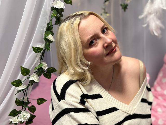 VictoriaNotti with blonde hair, smiling, wearing a white and black striped sweater, off-shoulder, cute, light skin, subtle makeup, pink pillow with white dots in the background.