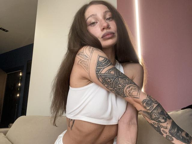 HotCaramelsTrio with long brown hair, nose ring, and tattoos on arms, wearing a white ribbed crop top, exposing a flat, toned stomach, and small, perky tits.