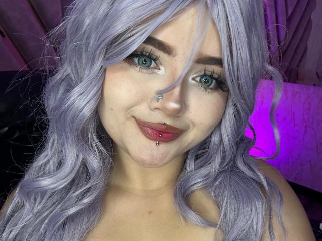 Lyanekkoo has purple wavy hair, blue eyes, a nose piercing, and a lip piercing with a ball. She's smiling with red lipstick. Her tits are not visible.