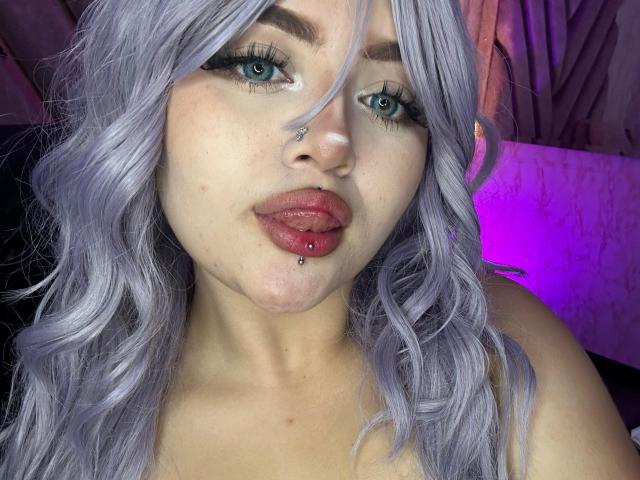 Lyanekkoo has pale purple wavy hair, blue eyes, black eyeliner, and a nose ring. She's pouting with a lip ring and a small facial mole.