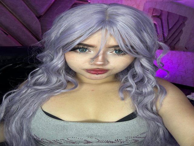 Lyanekkoo with long, wavy, silver hair, blue eyes, nose ring, red lipstick, and a grey tank top showing her small tits. She's staring at the camera with a neutral expression.