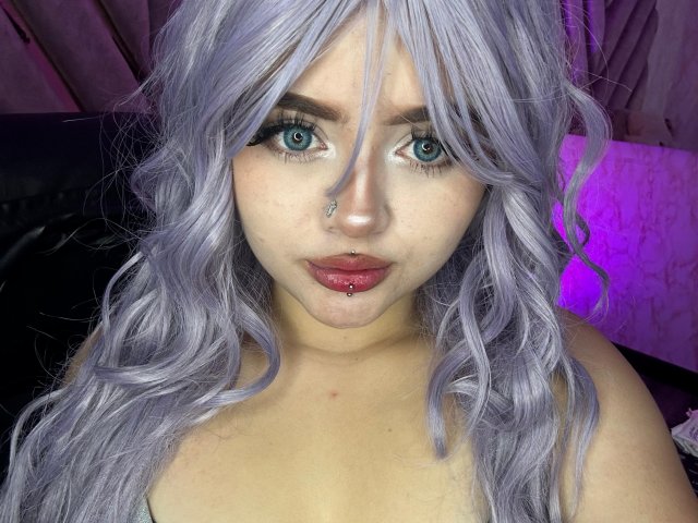 Lyanekkoo has pale skin, blue eyes, and wavy, silver hair. She has a nose piercing, lip piercing, and is looking at the camera with a serious expression.
