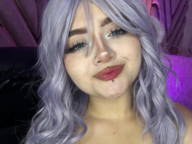 Lyanekkoo with light purple wavy hair, blue eyes, nose piercing, lip piercing, and red lipstick, smiling at the camera. She's got long lashes and a cute face.