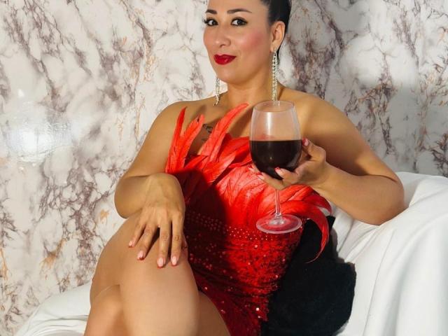 Karimelee with red sequined dress, holding glass of red wine, sitting on white chair, wearing earrings, black hair, red lipstick, black tattoo on chest, marble wall background.