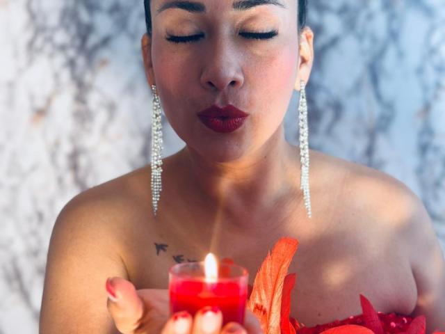 Karimelee with closed eyes, red lipstick, and diamond earrings, blowing out a red candle flame, holding red leaves, showing small tits, tattoo on left shoulder.