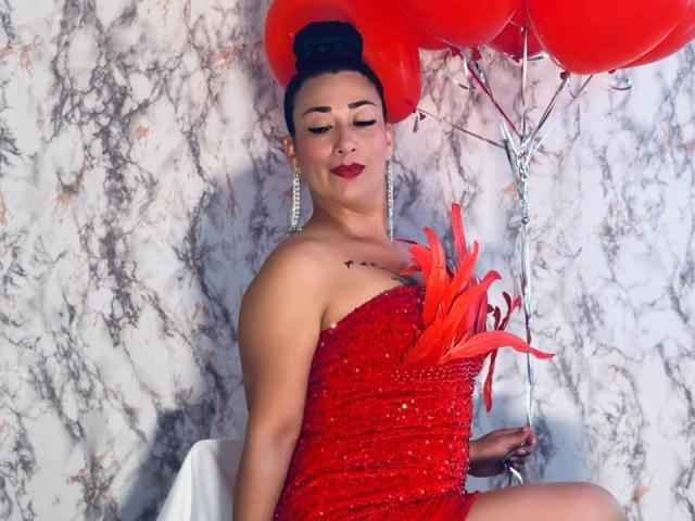 Karimelee with red sequined dress, holding red balloons, sitting with red feathers on her chest, wearing red lipstick, and earrings, dark hair in an updo, eyes closed.