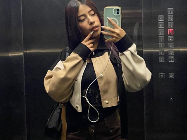 ScarletLuv, met lang donker haar, draagt een beige en zwarte jas, een zwart shirt en bruine broek, en maakt een selfie in een lift, met oordopjes en een zwarte tas..