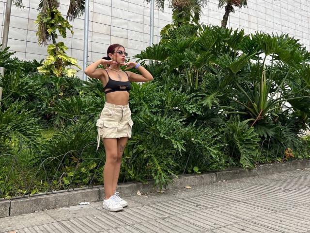 ScarletLuv met kleine borsten, een bruine bh, een beige rok, witte sneakers, donker haar, een zonnebril, met een telefoon aan haar oor, staand op de stoep, met groene planten, een grijze betegelde muur op de achtergrond..