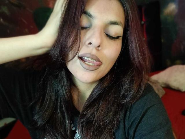 SoyVeronica has dark brown hair, closed eyes with long eyelashes, and is sticking out her tongue while touching her head with a hand. She's wearing a black shirt.